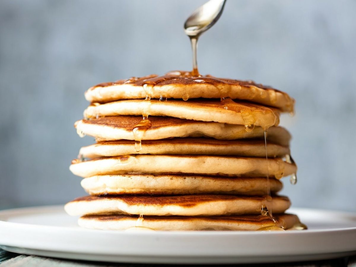 Ein Teller mit einigen gestapelten Pancakes, darüber ein Löffel, der Honig darüber gibt.