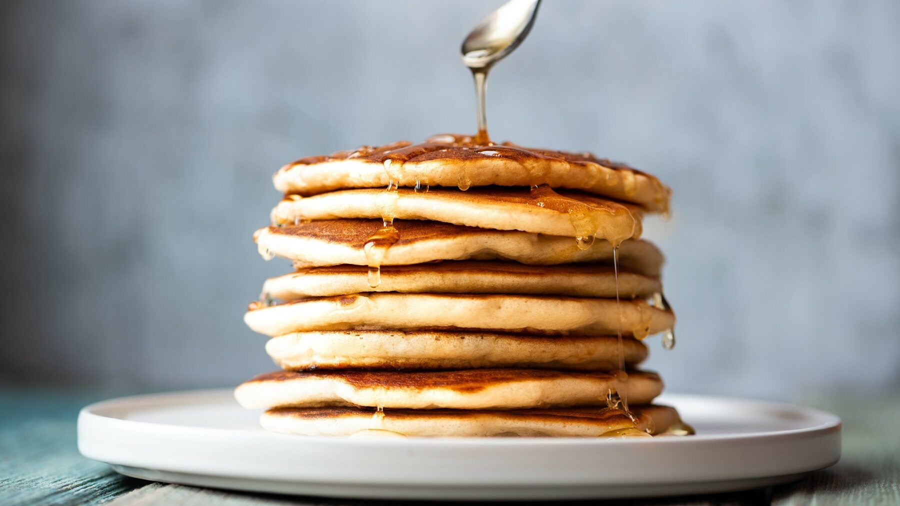 Ein Teller mit einigen gestapelten Pancakes, darüber ein Löffel, der Honig darüber gibt.
