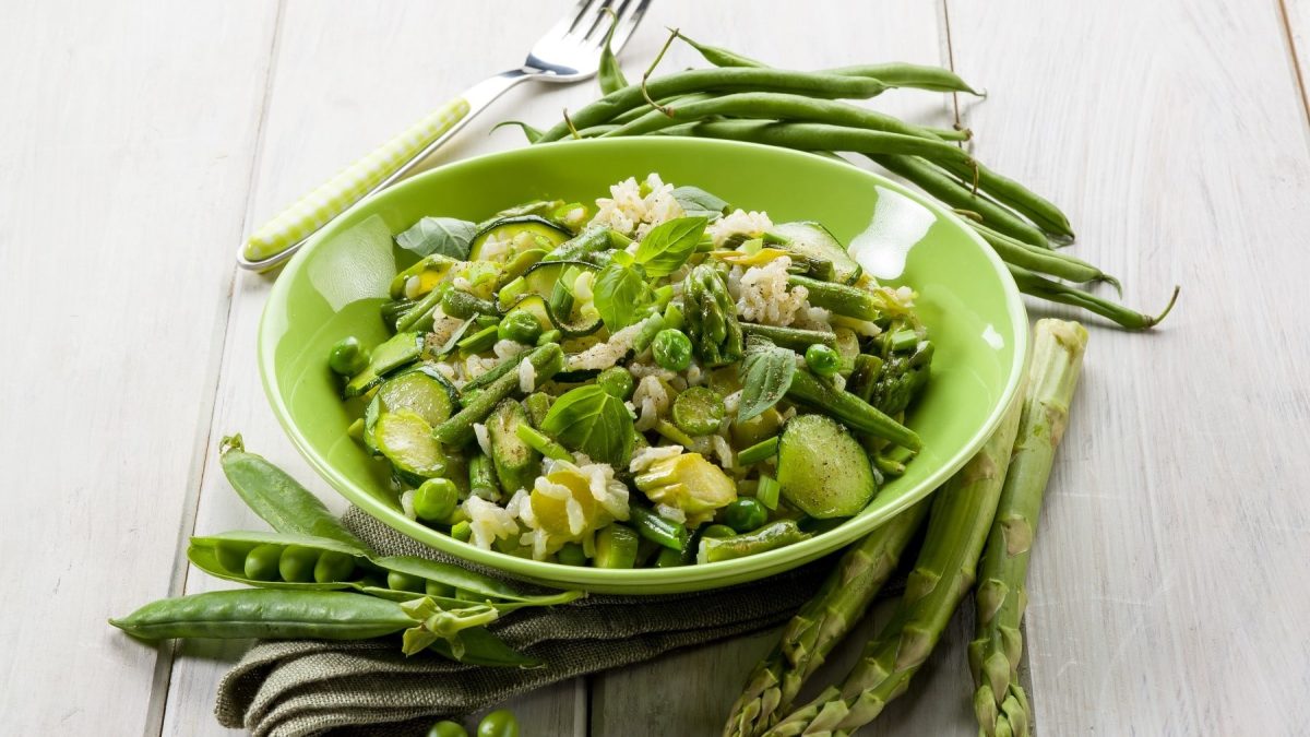 Grünes Spargel-Risotto mit Zucchini, Erbsen, Frühlingszwiebeln und frischem Basilikum, serviert in einer grünen Schale auf einem hellen Holztisch.