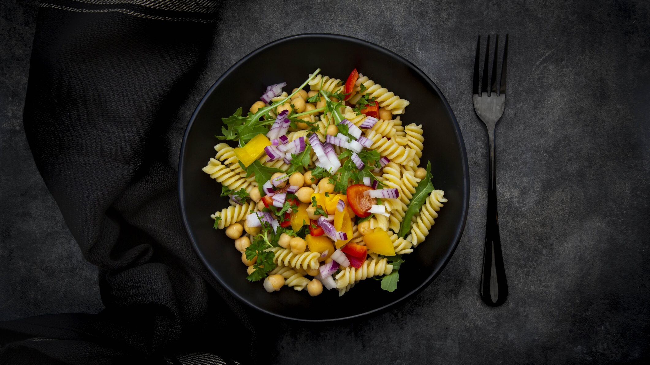 Rucola-Nudel-Salat mit Kichererbsen in schwarzer Schüssel, daneben Gabel