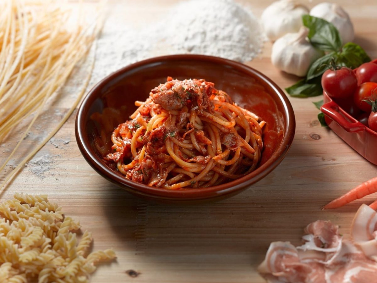 In einer Schale angerichtete Spaghetti-Pasta mit geräuchertem Schinken aus Parma und Tomatensauce, umgeben von den Zutaten der Sauce wie Knoblauch, frischen Tomaten und dem Parmaschinken.