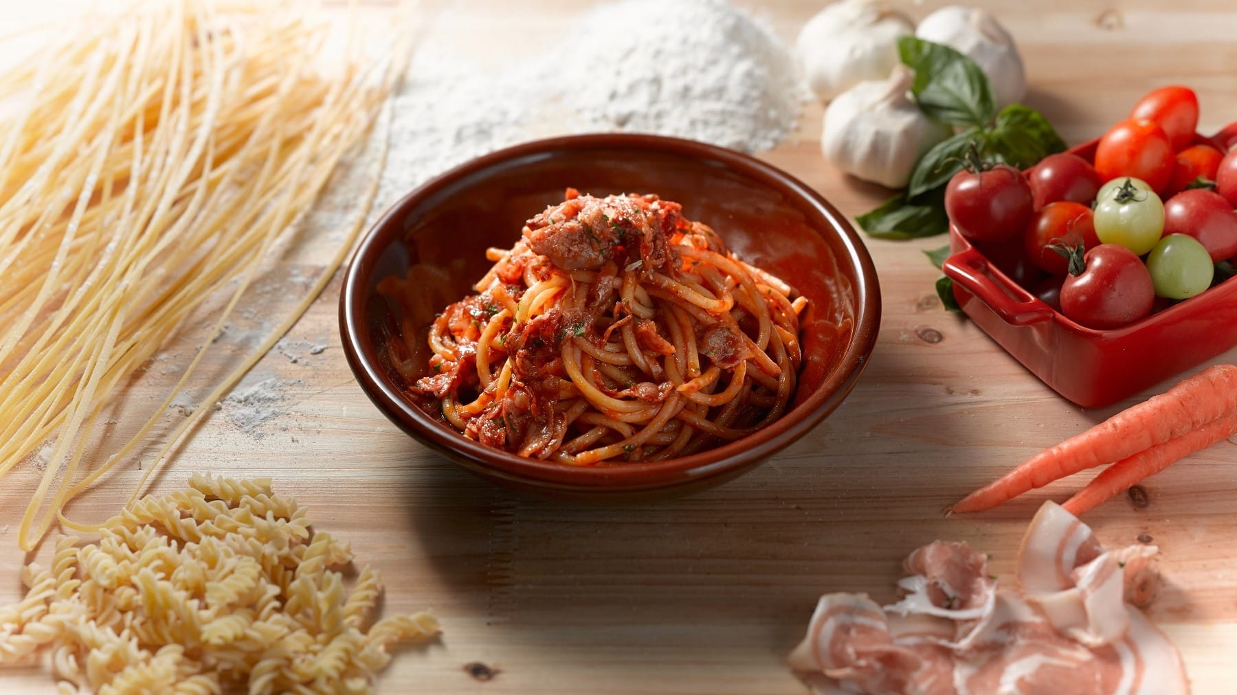 In einer Schale angerichtete Spaghetti-Pasta mit geräuchertem Schinken aus Parma und Tomatensauce, umgeben von den Zutaten der Sauce wie Knoblauch, frischen Tomaten und dem Parmaschinken.
