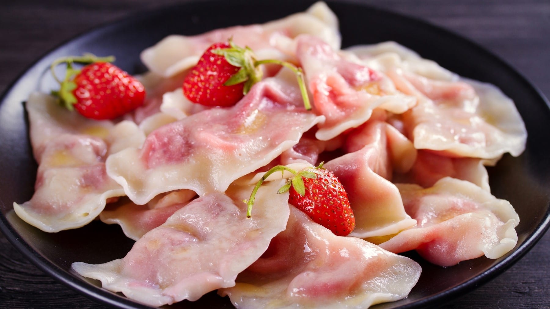 Fruchtig gefüllte Pierogi mit Erdbeerfüllung, ausgekocht und serviert auf einem dunklen Teller, garniert mit frischen Erdbeeren.