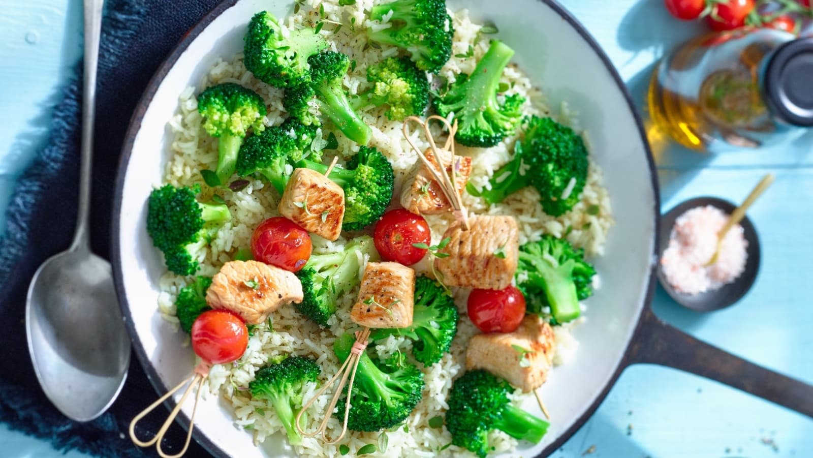 Draufsicht: Blauer Untergrund mit einer Pfanne in der Bildmitte. Darin weißer Reis mit Brokkoli und Schaschlikspießen aus Fleisch und frischen Tomaten.