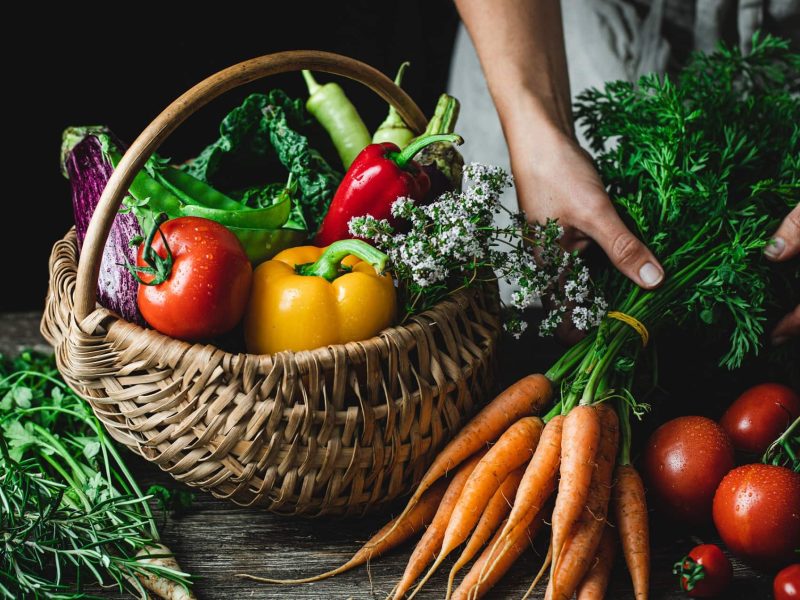 Clean Eating: ein Korb und Tisch mit Gemüse, ein Bund Karotten wird von einer Frau gehalten.