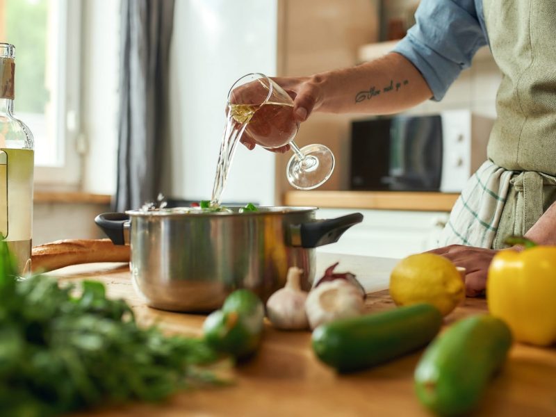 Mann mit Tattoo am Arm steht in einer hellen Küche und gießt Weißwein in einen Topf, der hinter Gemüse auf einer hellen Arbeitsplatte aus Holz steht.