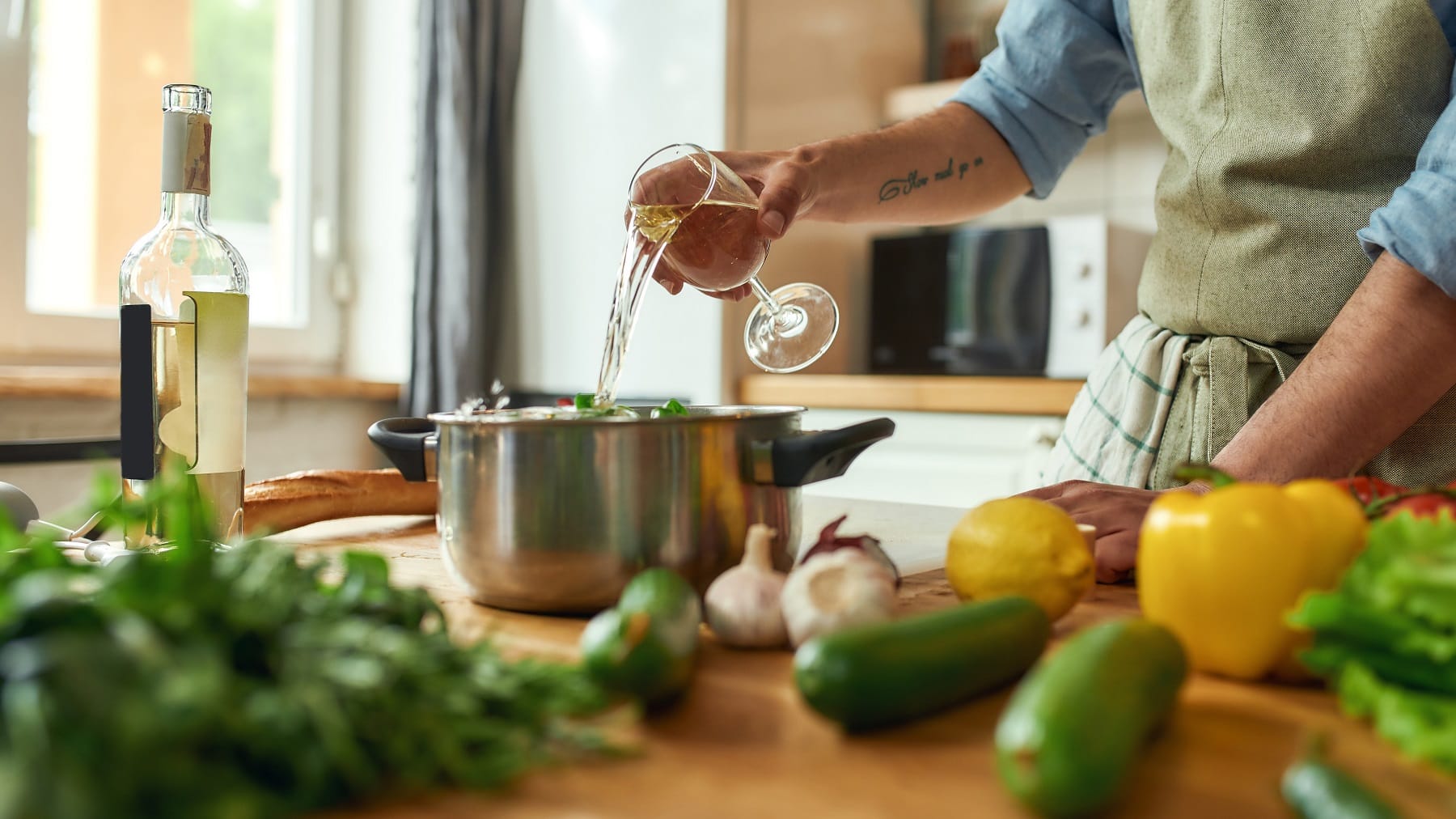 Mann mit Tattoo am Arm steht in einer hellen Küche und gießt Weißwein in einen Topf, der hinter Gemüse auf einer hellen Arbeitsplatte aus Holz steht.