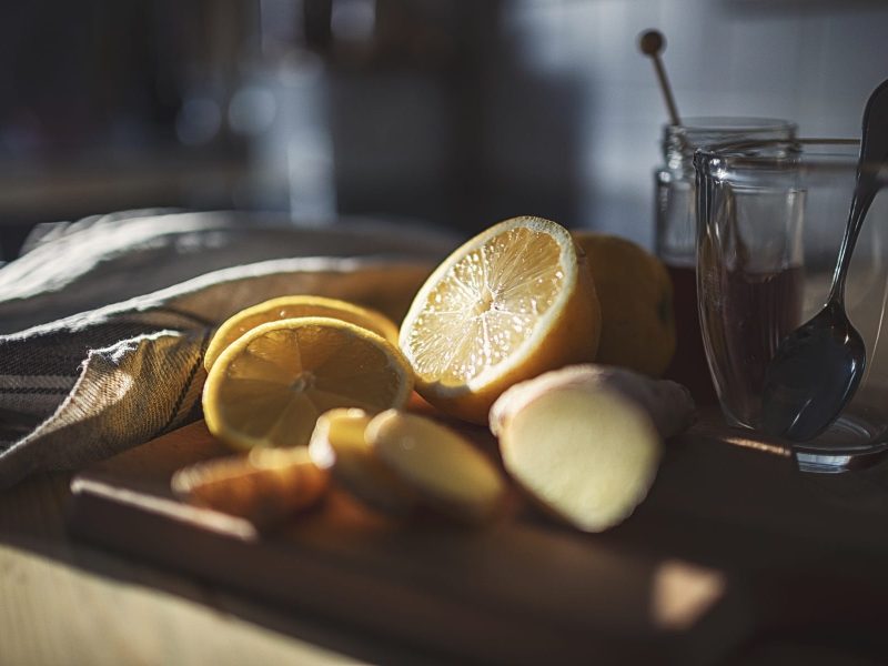 Nahaufnahme von aufgeschnittener Zitrone und Ingwer neben einem leeren Glas auf einem hellen Schneidebrett auf Holz und einem grauen Geschirrtuch.