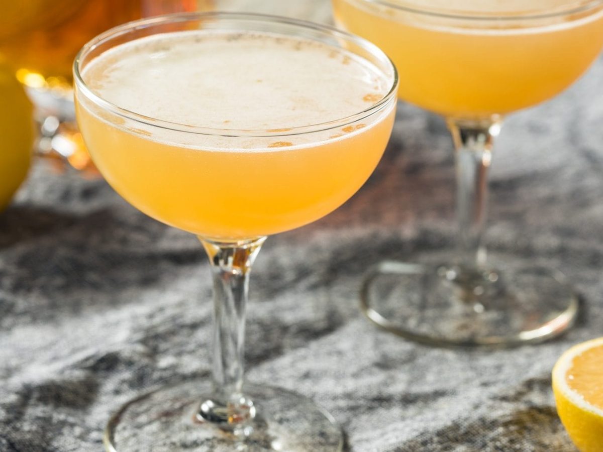 Zwei Gläser mit Champagner Fizz auf einem Tisch mit grauer Tischdecke, mit frischen Zitronen und einer Flasche Cognac im Hintergrund.