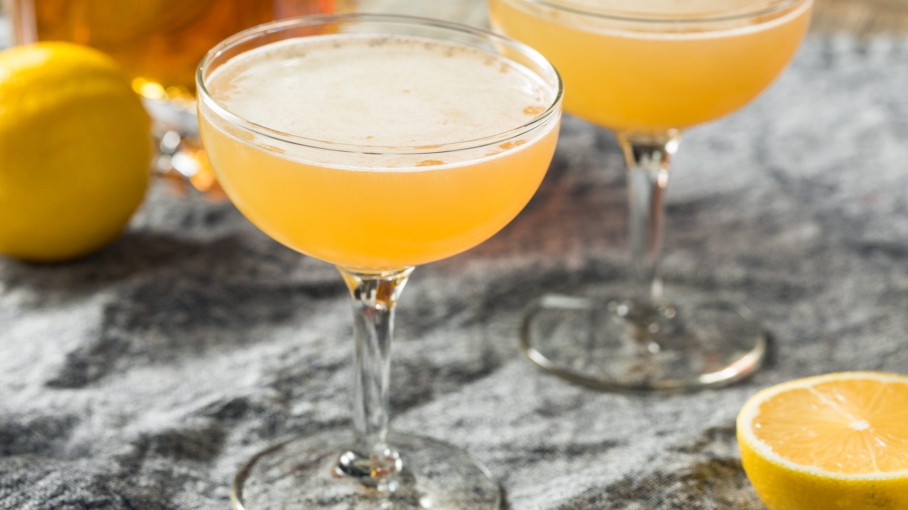 Zwei Gläser mit Champagner Fizz auf einem Tisch mit grauer Tischdecke, mit frischen Zitronen und einer Flasche Cognac im Hintergrund.