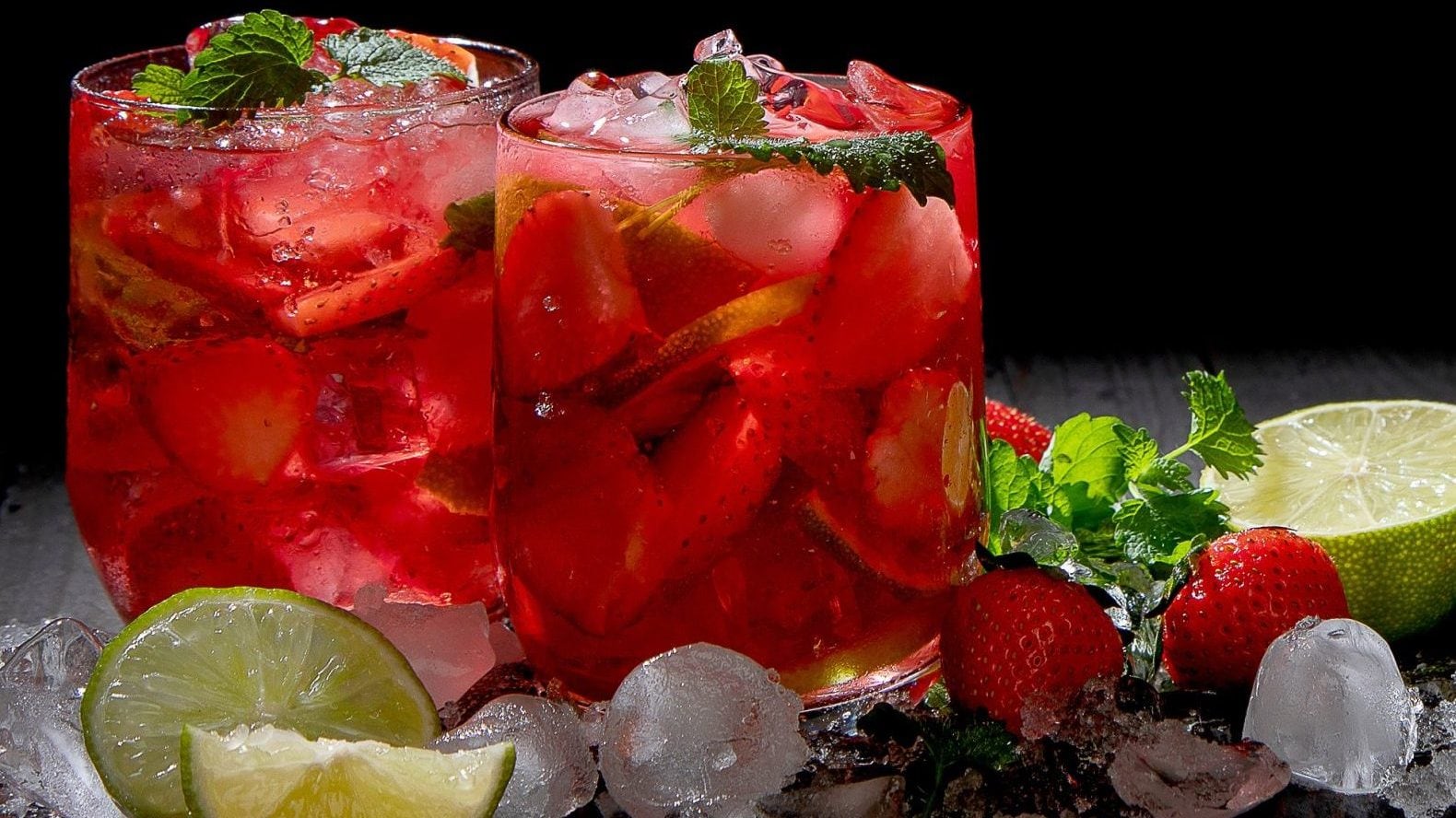 Erdbeer-Mojito in zwei Gläsern auf dunklem Untergrund, daneben Crushed Ice, Erdbeeren, Minze und Limetten.