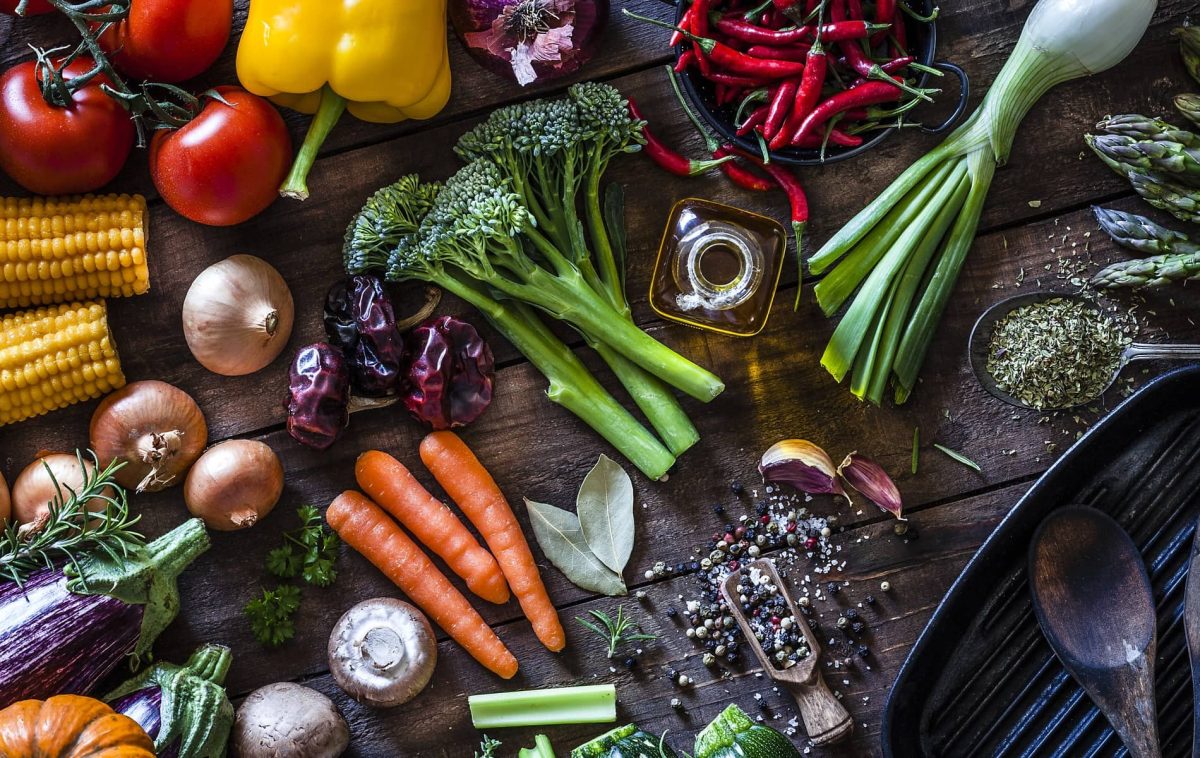 Verschiedene Lebensmittel, wie Brokkoli, Karotten, Mais, sowie weiteres Gemüse und verschiedene Kräuter und Würzmittel liegen auf einem Tisch verteilt.