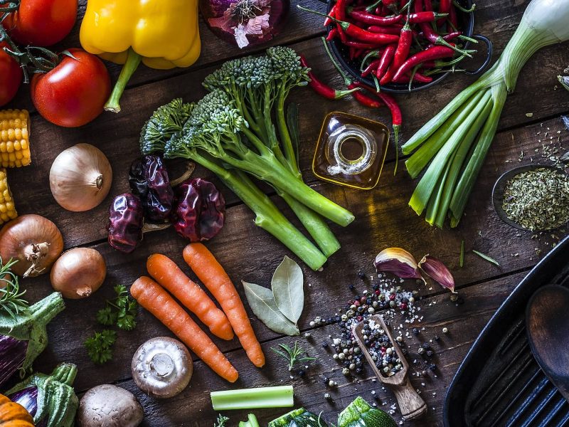 Verschiedene Lebensmittel, wie Brokkoli, Karotten, Mais, sowie weiteres Gemüse und verschiedene Kräuter und Würzmittel liegen auf einem Tisch verteilt.