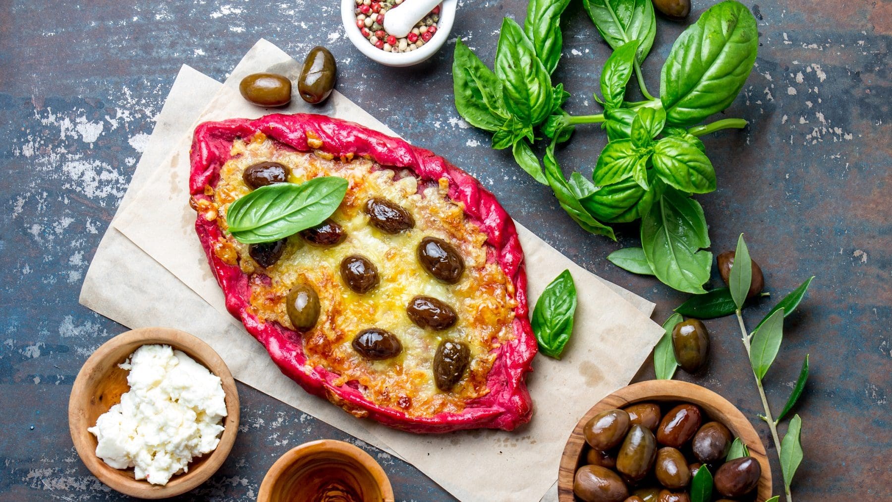 Eine ovale Pinke Pizza mit feta und Oliven liegt auf zwei hellbraunen Papierstreifen. Der Untergrund ist Metall mit Rost-Optik. Um die Pizza herum stehen Schüsseln mit Feta, Oliven und Pfeffer. Auf dem Untergrund verteilt sind Basilikum und Oliven.