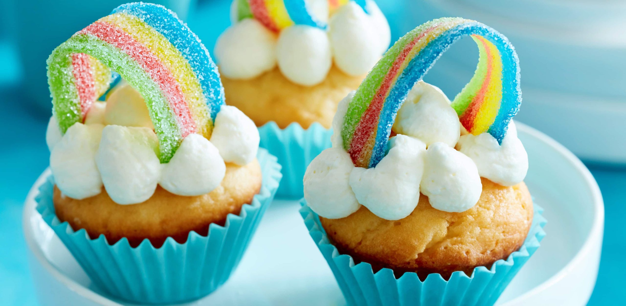 Rainbow-cupcakes in blauen Förmchen mit Regenbogen und Sahne auf einem weißen Ständer.
