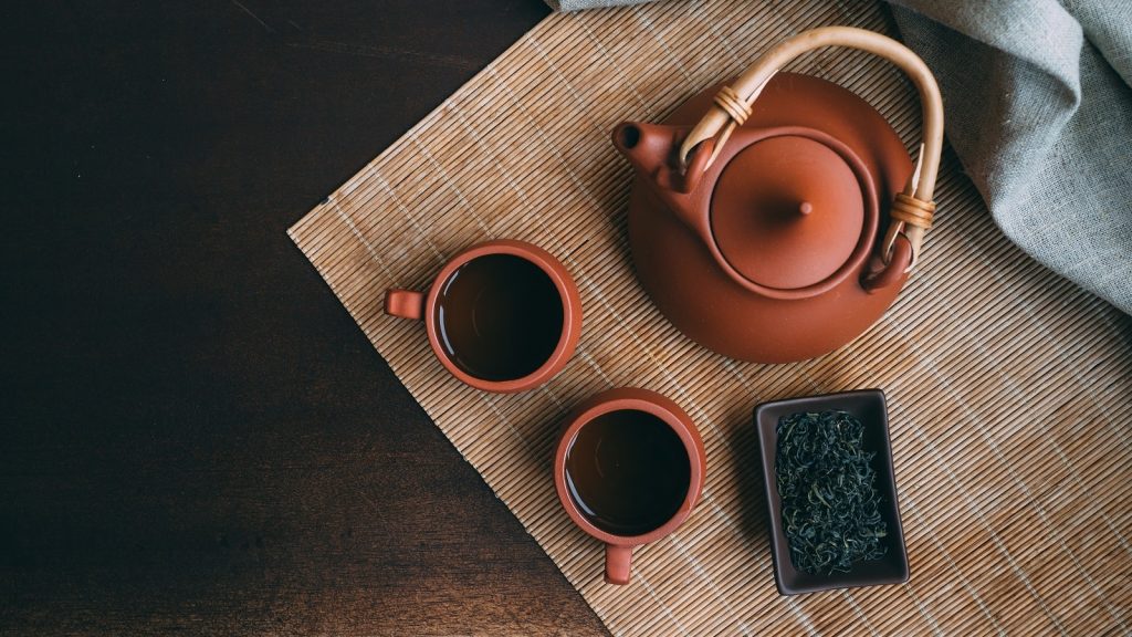 Eine Ton-Teekanne neben zwei Ton-Tassen mit Grünem Tee auf einer Bambus Unterlage von oben.