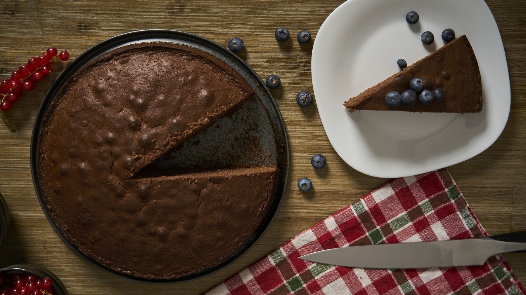 Ein angeschnittener Depression-Cake auf einem dunklen Kuchenteller steht auf einem braunen Tisch. Rechts neben dem Kuchen steht ein weißer Teller mit einem Stück des Kuchens. unter dem Teller liegt ein kariertes Handtuch mit einem Kuchenmesser.