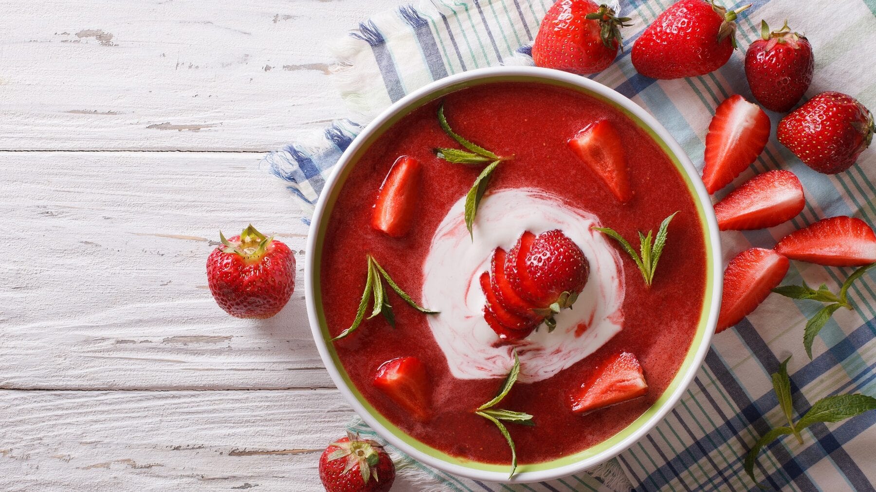 Erdbeersuppe mit frischen Erdbeeren, Quark und Minze in einer weißen Schüssel auf einem weißen Tisch mit einem Geschirrtuch und frischen Erdbeeren.