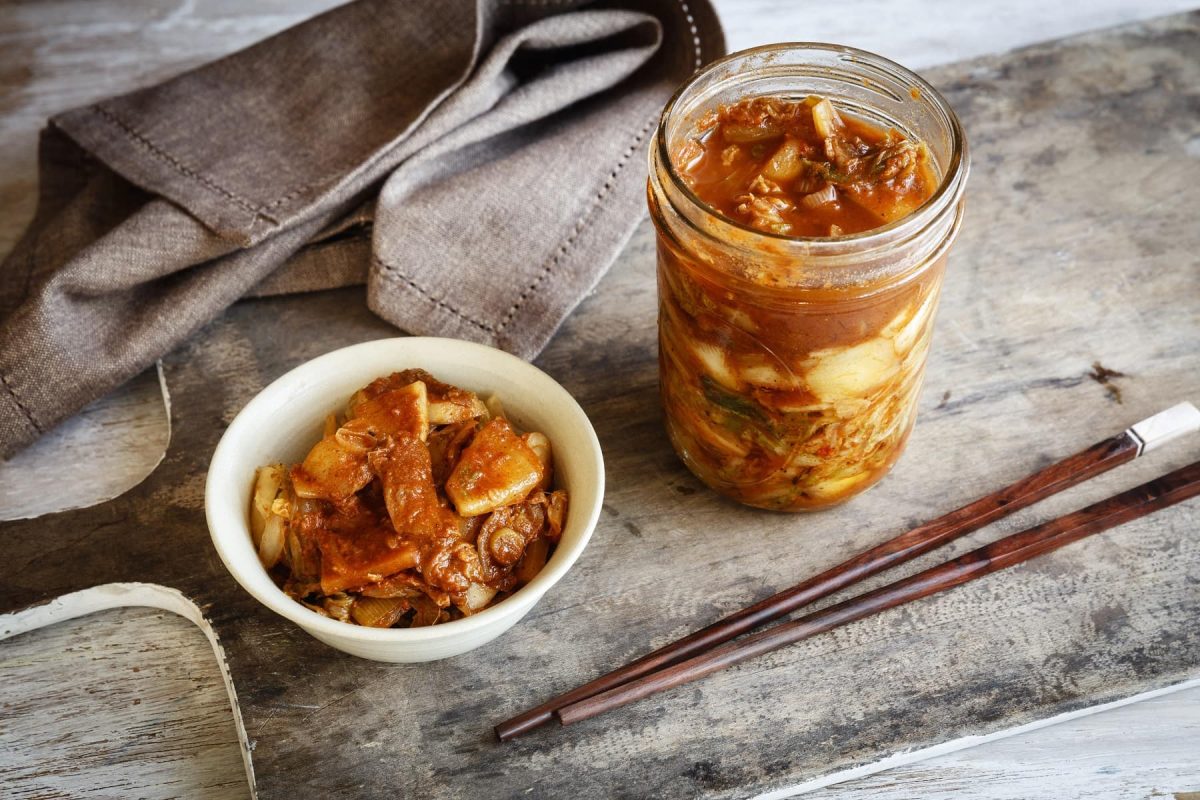 Ein Glas mit fermentiertem Kimchi steht neben einer Schale Reis und Kimchi, daneben liegen Stäbchen.