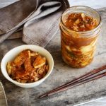 Ein Glas mit fermentiertem Kimchi steht neben einer Schale Reis und Kimchi, daneben liegen Stäbchen.