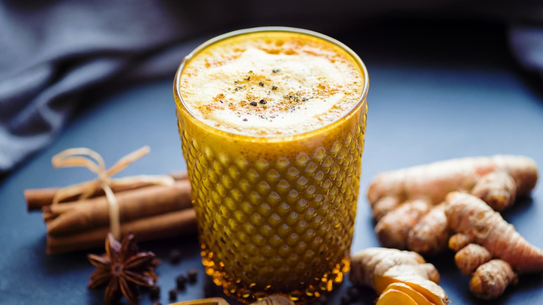Goldene Milch in einem Glas auf einer Schieferplatte neben Kurkuma, Zimt und Anis vor einem dunklen Tuch.