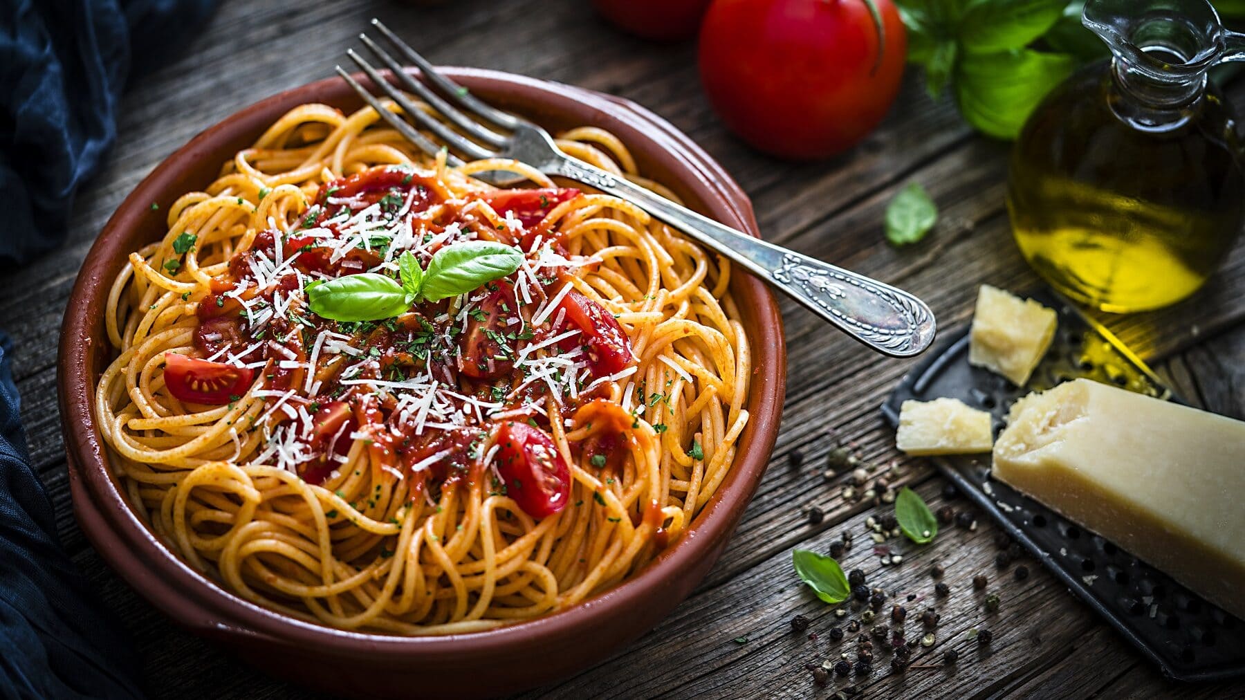 Spaghetti-Salat mit Tomatensoße, frischem Basilikum und geriebenem Parmesan in einer braunen Tonschüssel auf einem dunklen Holztisch.