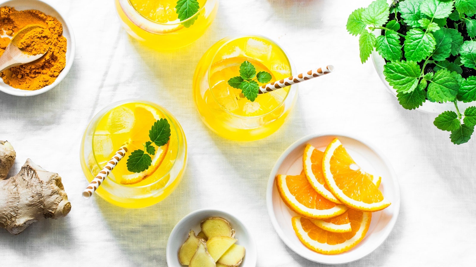 Zwei Gläser gefüllt mit Limonade mit Kurkuma und Inger und garniert mit Orangenscheiben und Minze, neben einer Schale Kurkuma-Pulver, Ingwerscheiben und Orangenscheiben, von oben.