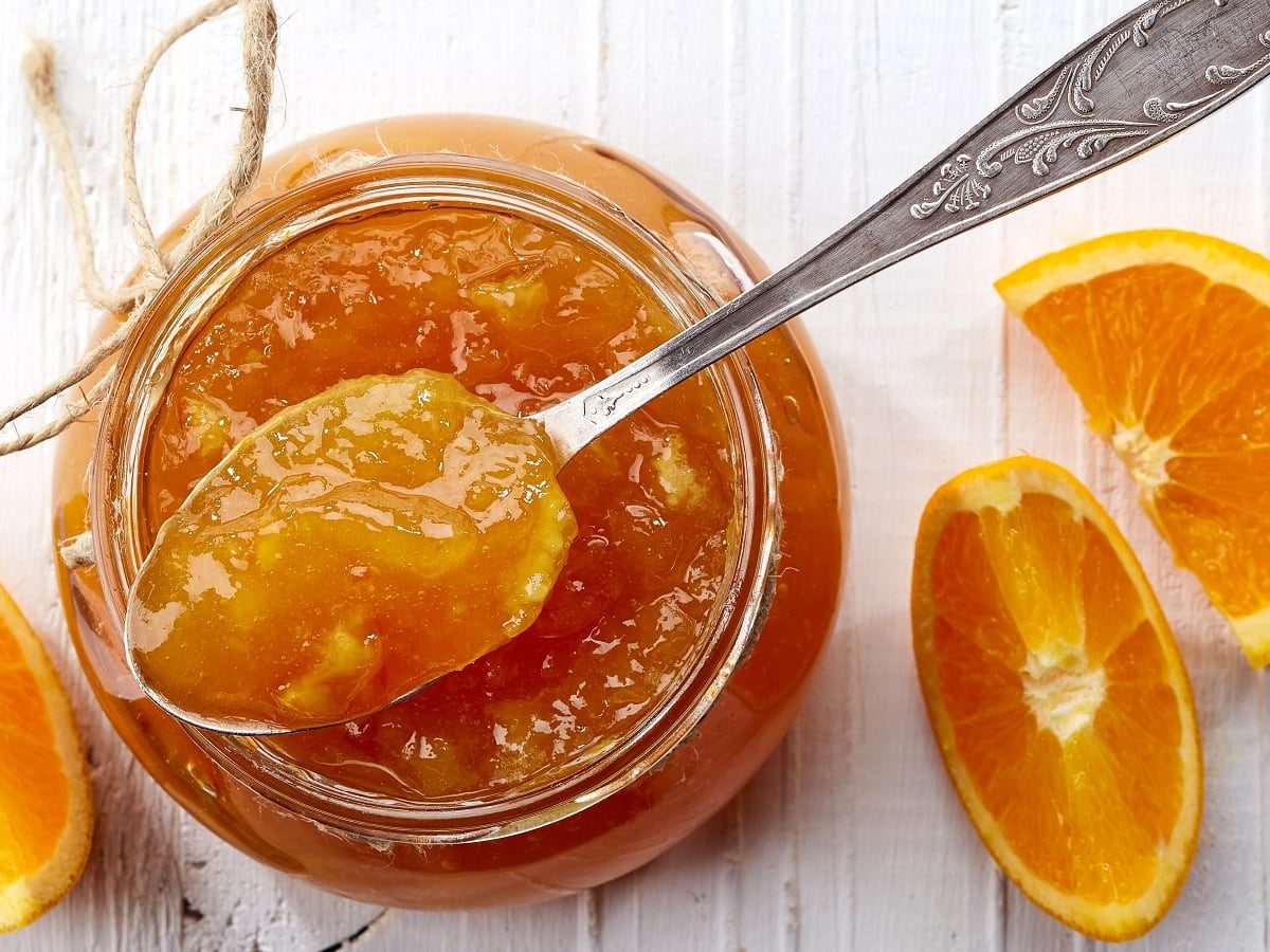 Ein offenes Glas Orangen-Grapefruit-Marmelade mit Schuss. Ein Teelöffel entnimmt dem Glas ein wenig der Marmelade. Orangen-Scheiben liegen zur Dekoration neben dem Glas, alles von oben fotografiert.