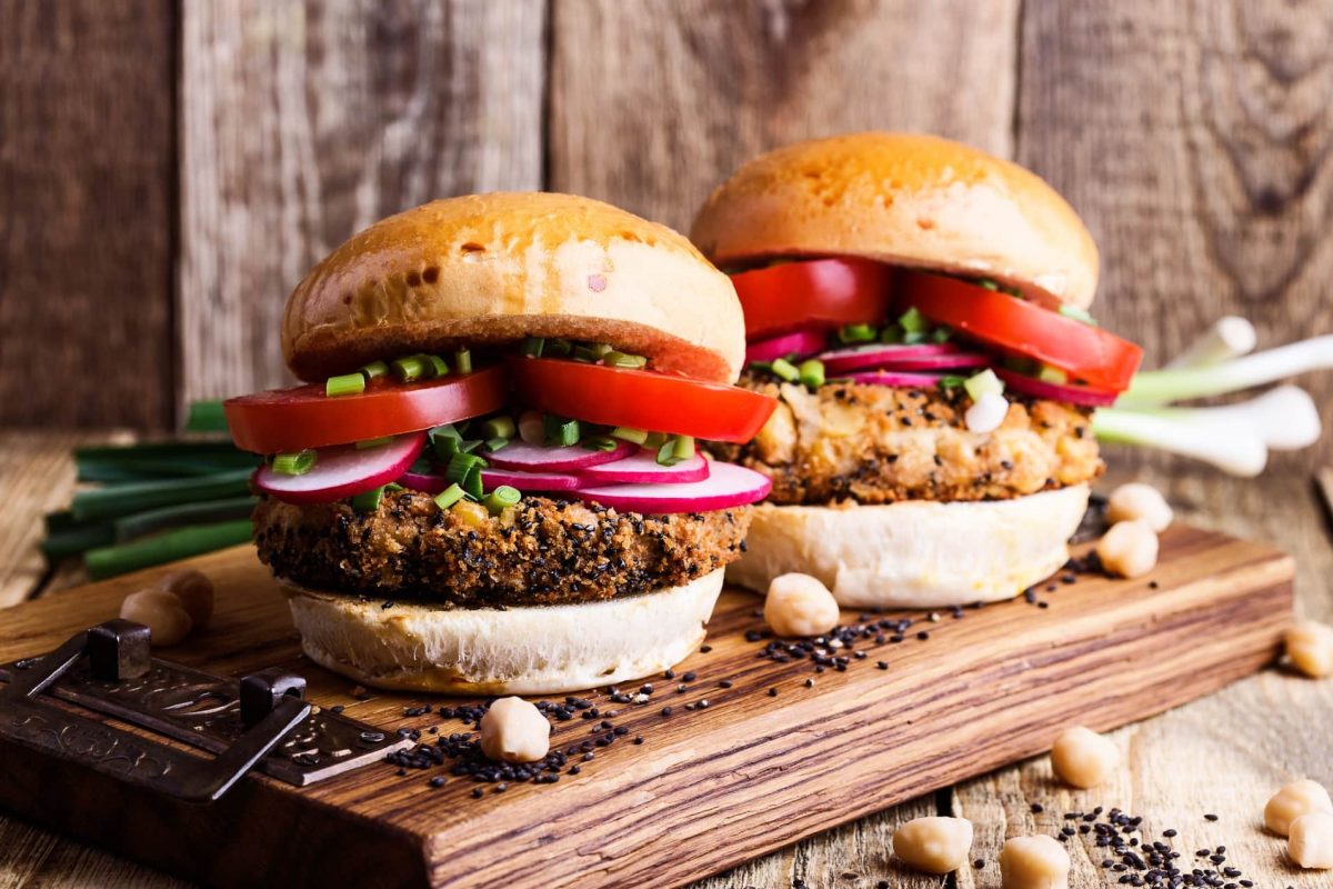 Herstellung von Fleischersatz-Produkten stark gestiegen: Zwei üppig belegte Veggie-Burger auf einem Brett, daneben einige Kichererbsen.