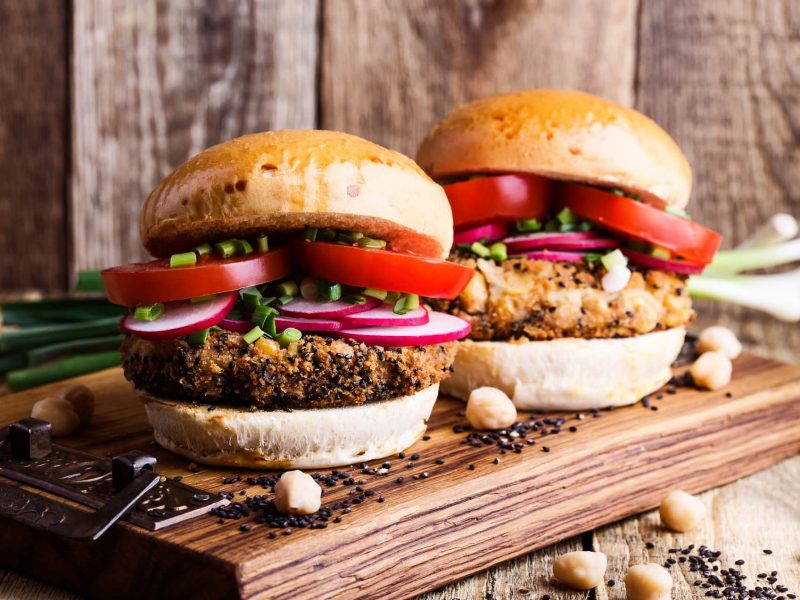 Herstellung von Fleischersatz-Produkten stark gestiegen: Zwei üppig belegte Veggie-Burger auf einem Brett, daneben einige Kichererbsen.