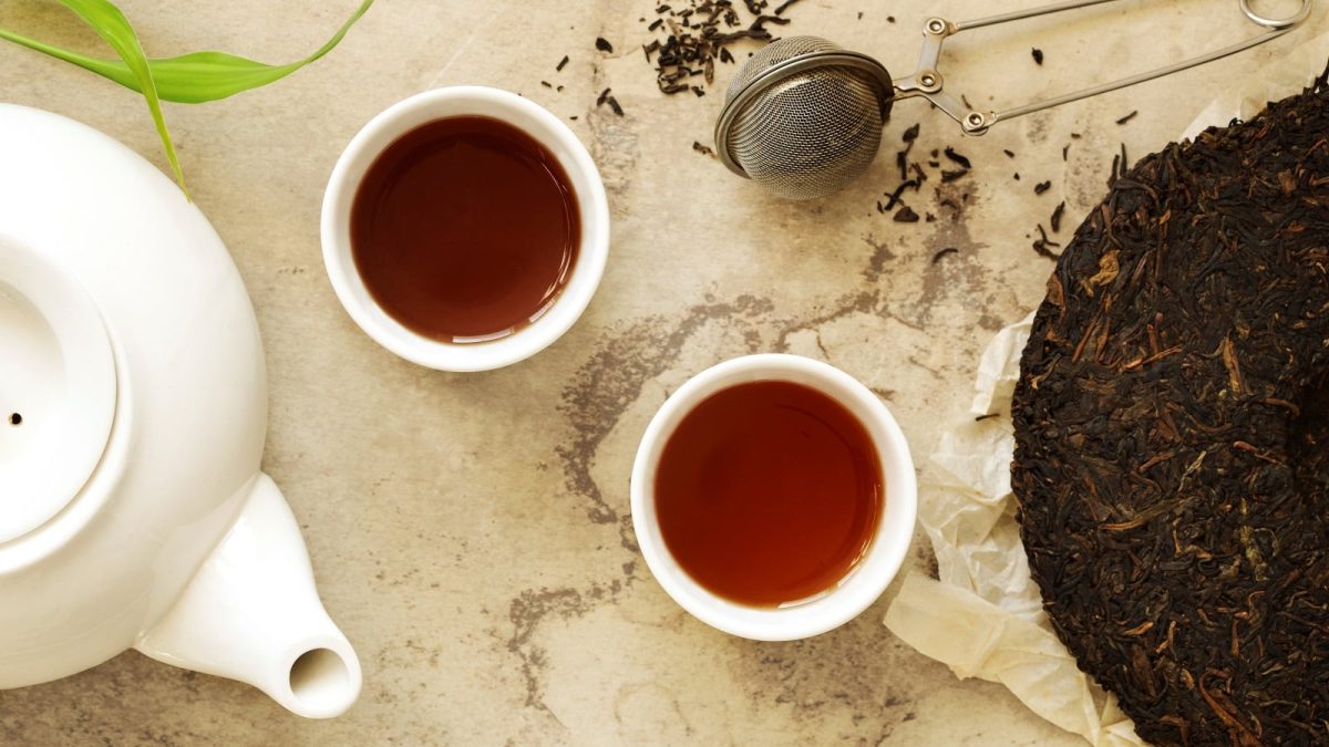 Zwei weiße Tassen mit dunklem Pu Erh-Tee neben einer weißen Teetasse und einem Stück dunklen Pu Erh Tee-Kuchen, vor einem Marmorhintergrund, von oben.