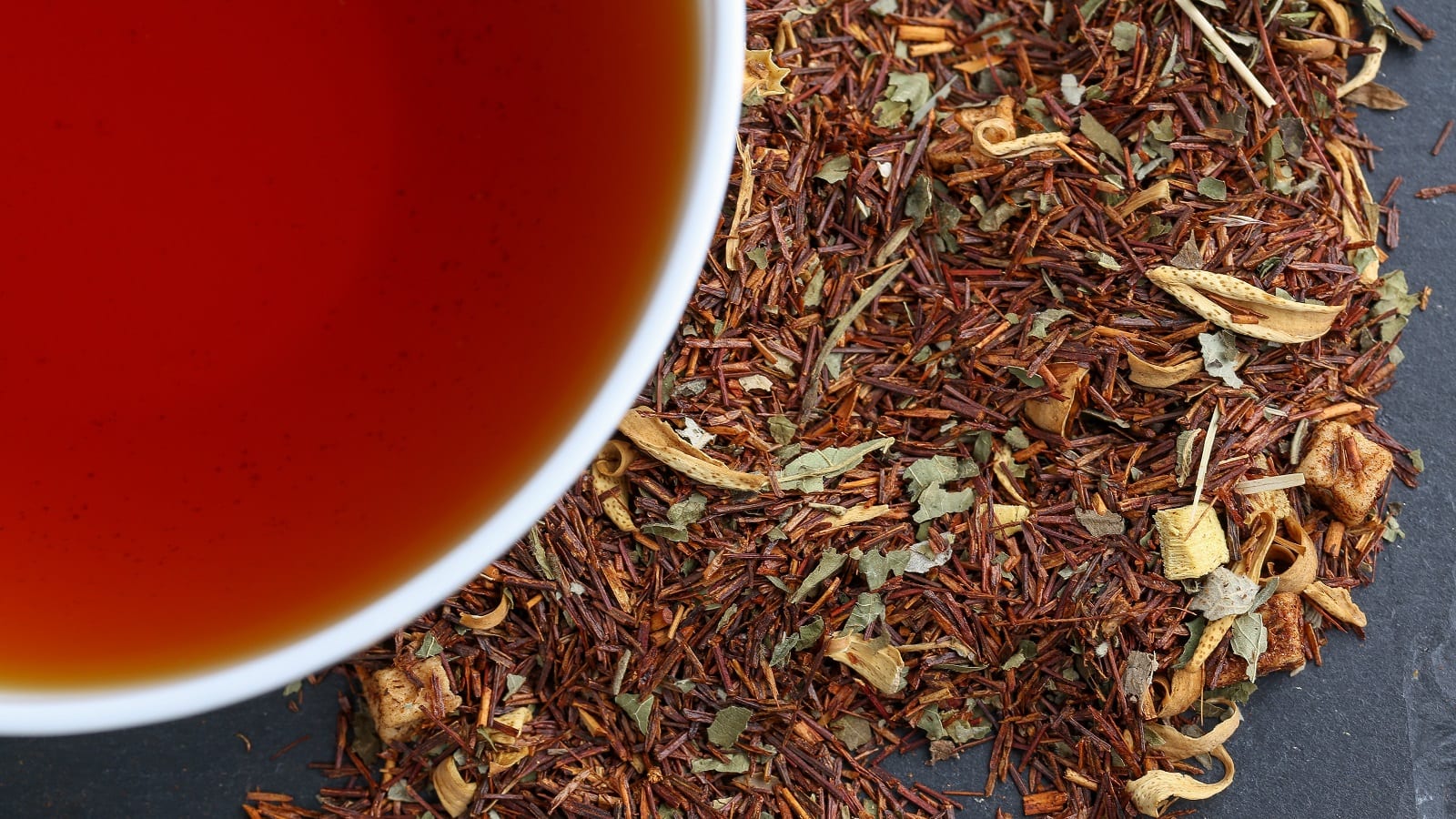 Eine weiße Tasse roter Rooibostee neben getrockneten Teeblättern.