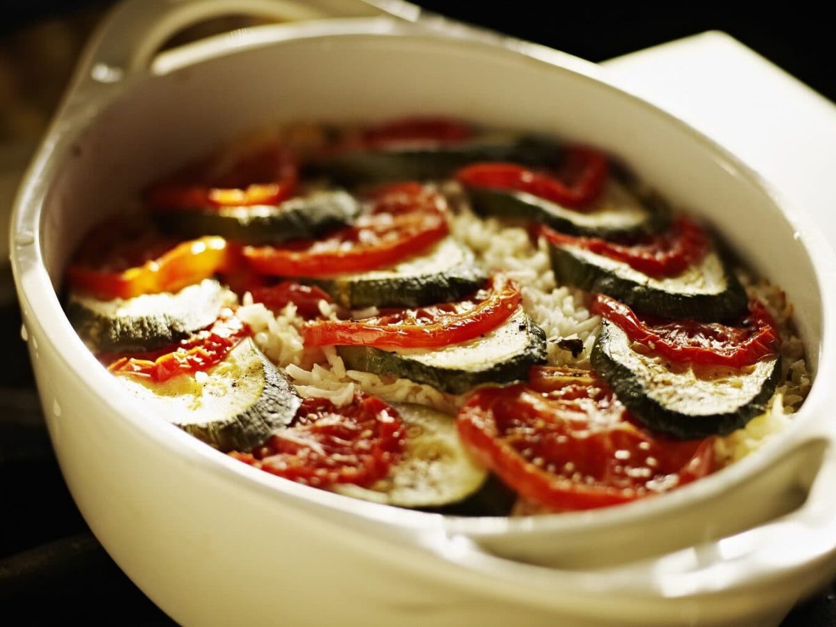 Zucchini Auflauf mit Tomate in einer hellen Auflaufform mit Parmesan überbacken auf einem dunklen Tisch mit Sonne von hinten