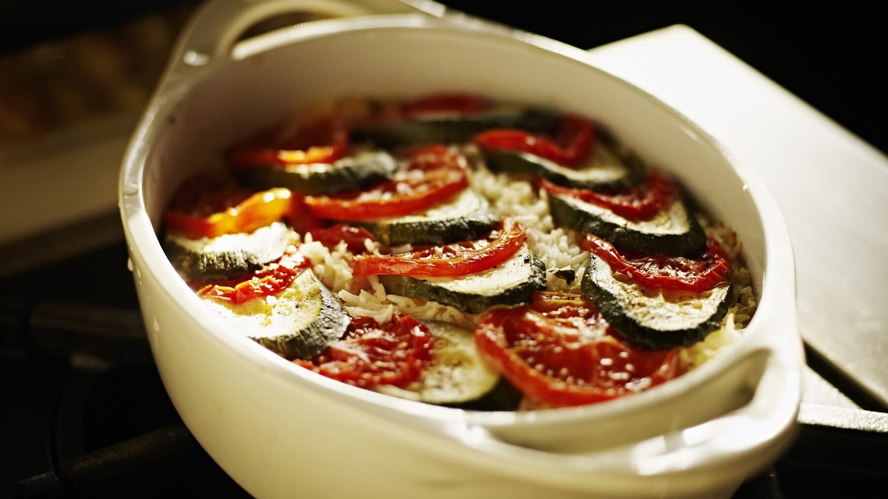 Zucchini Auflauf mit Tomate in einer hellen Auflaufform mit Parmesan überbacken auf einem dunklen Tisch mit Sonne von hinten
