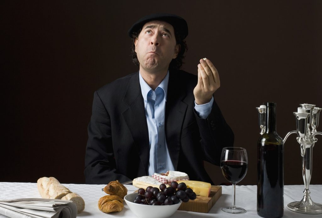 Ein Mann mit Hut und Sakko sitz an einem Tisch mit Käse und Wein und macht eine Geste zum Himmel