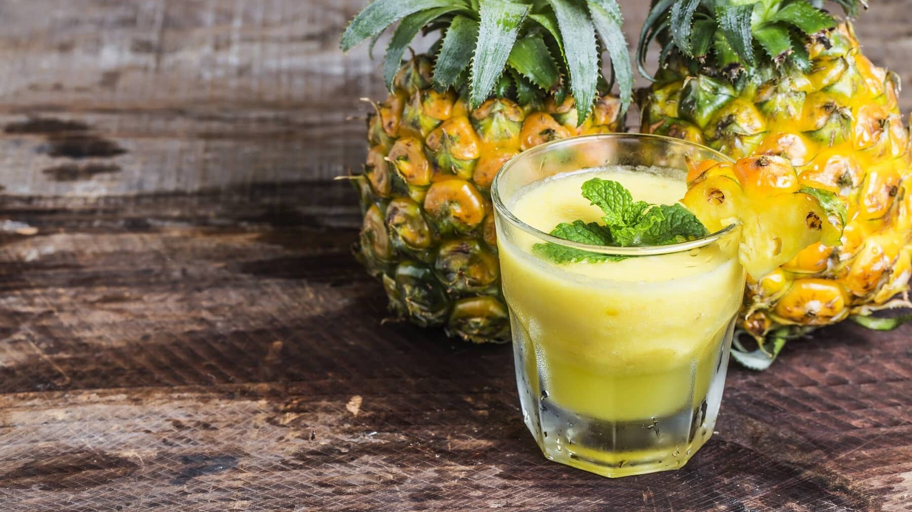 Ein Ananas-Smoothie in einem klekinen Glas steht auf einem Holztisch. Hinter dem Glas stehen zwei Ananasfrüchte. Der Smoothie ist angerichtet mit einer Scheibe Ananas und etwas Minze.