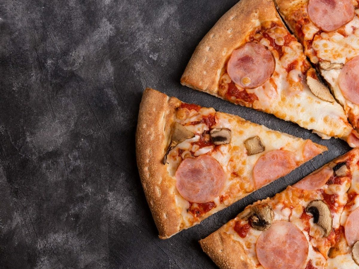Eine Champinon-Pizza mit Speck liegt auf einem dunklen Untergrund aus Stein. Die Pizza ist bereits angeschnitten und ein Stück steht ein Wenig hervor.