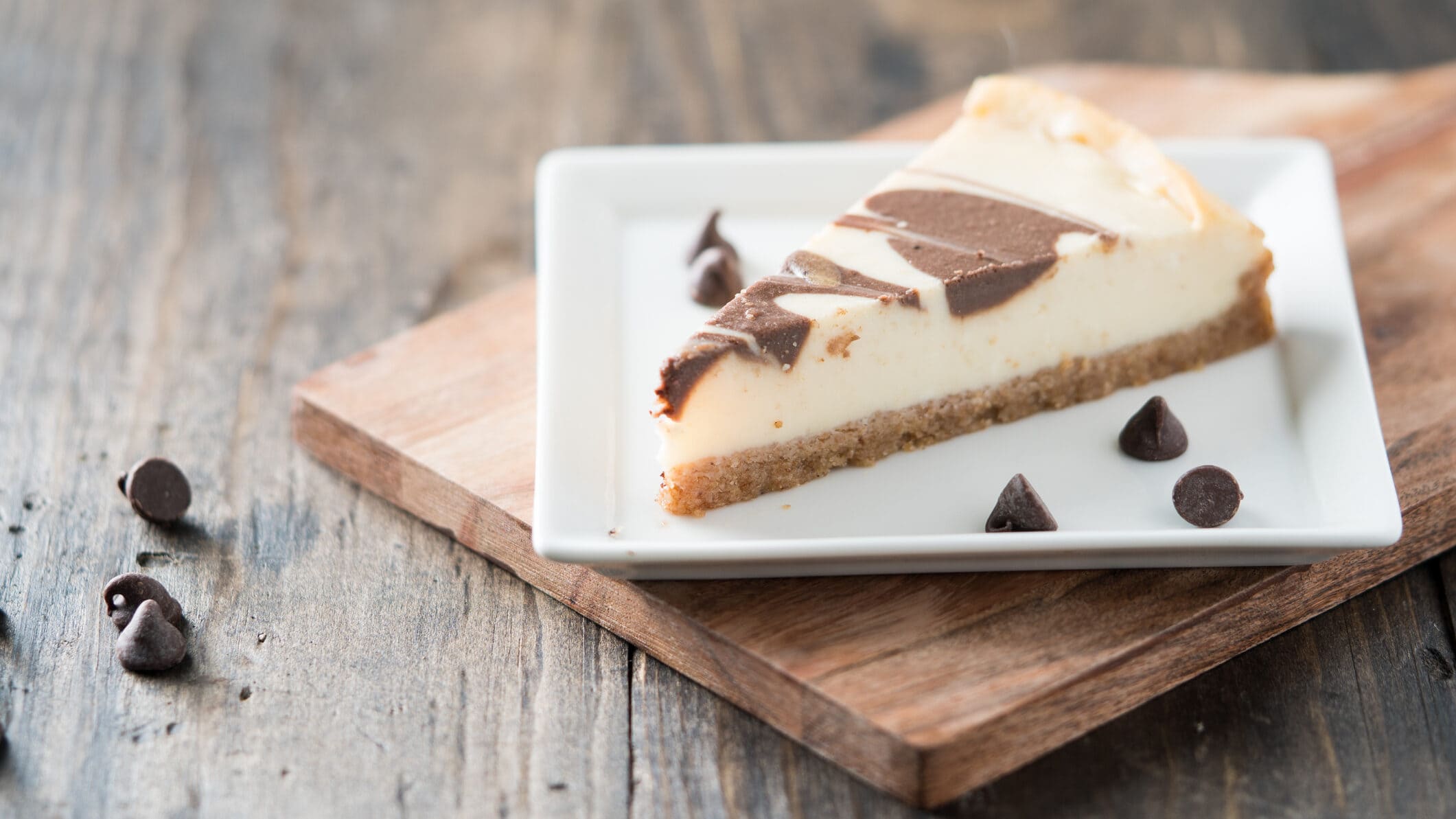 Ein Stück Cheesecake mit Schoko-Swirl auf einem Desserteller, der auf einem Holzbrett steht. Daneben ein paar Schoko-Drops