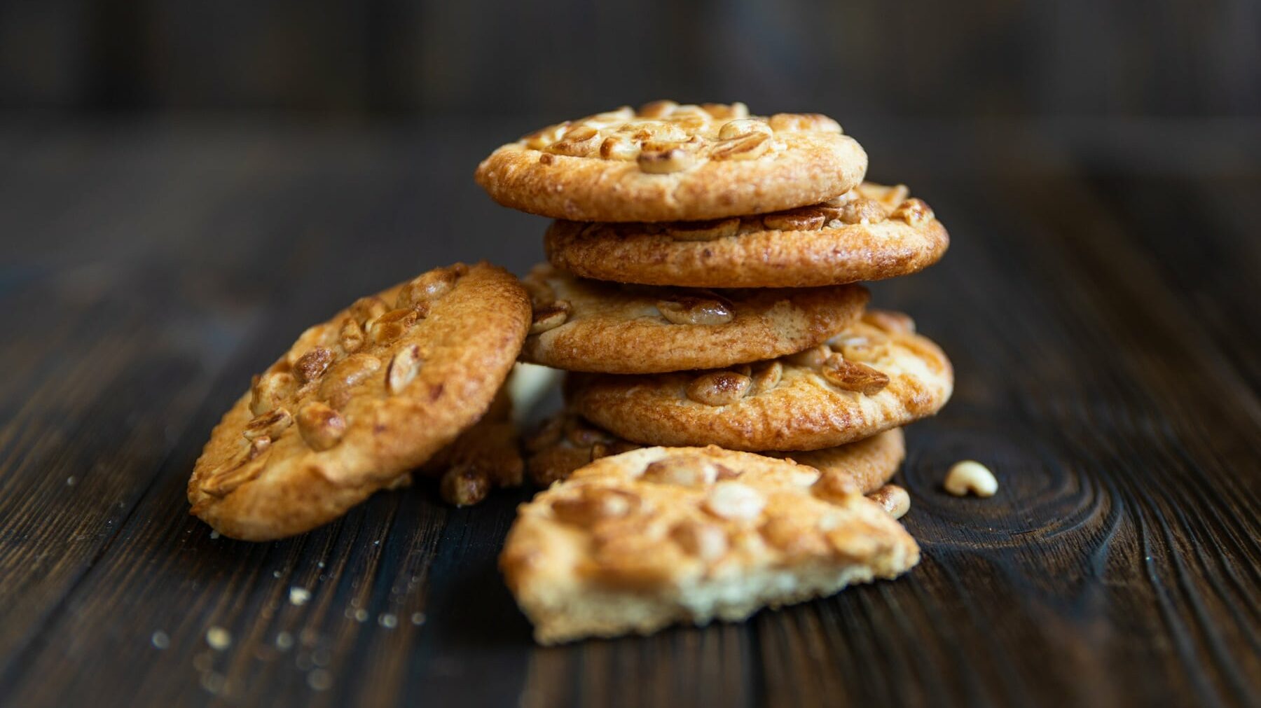 Erdnuss Cookies liegen gestapelt auf einem braunen Holztisch. Fünf Kekse bilden einen Turm, ein sechster Keks ist seitlich gegen den Turm gelehnt. Im Vordergrund liegt ein halbierter Keks und einige Krümel.