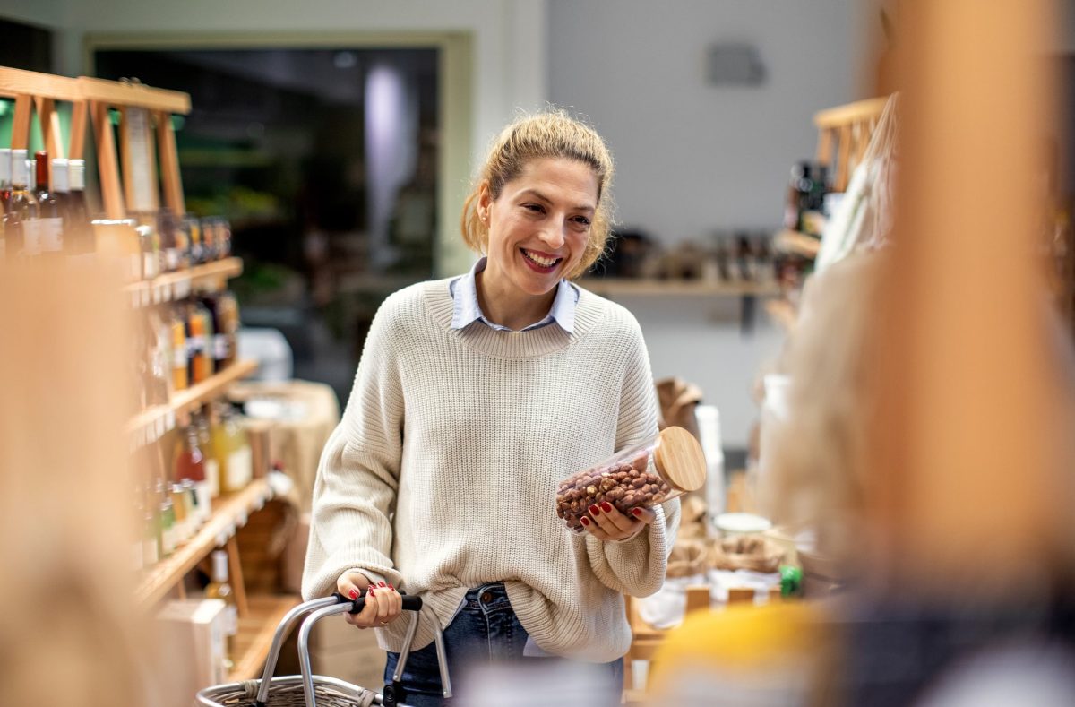 Foodhub München: Eine Frau steht in einem kleinen Supermarkt, lächelt und hält einen Einkaufswagen in der einen und eine Glasdose mit Nüssen in der anderen Hand.
