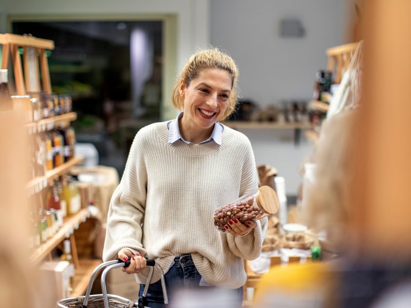 Foodhub München: Eine Frau steht in einem kleinen Supermarkt, lächelt und hält einen Einkaufswagen in der einen und eine Glasdose mit Nüssen in der anderen Hand.