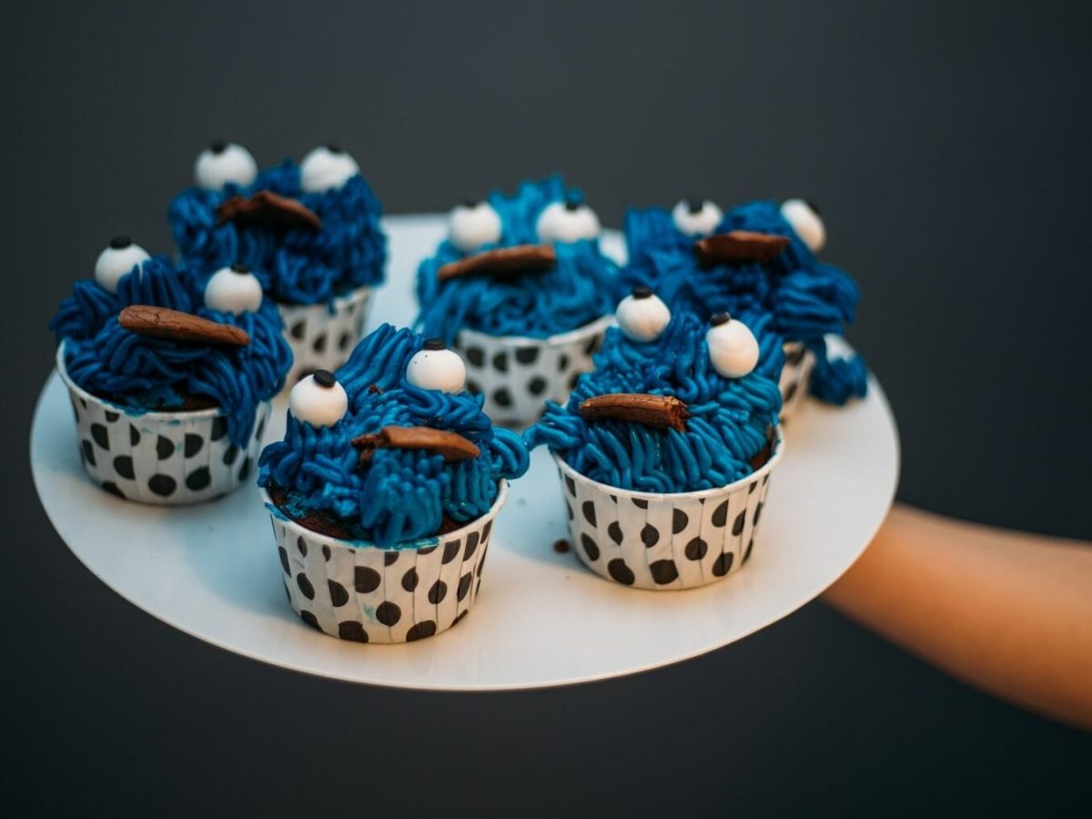Fünf Krümelmonster-Cupcakes stehen auf einem weißen Teller und werden vor einen schwarzen Hintergrund gehalten. Die Cupcakes stehen in gepunkteten Muffinformen und in jedem Krümelmonster-Cupcake steckt ein Keks.