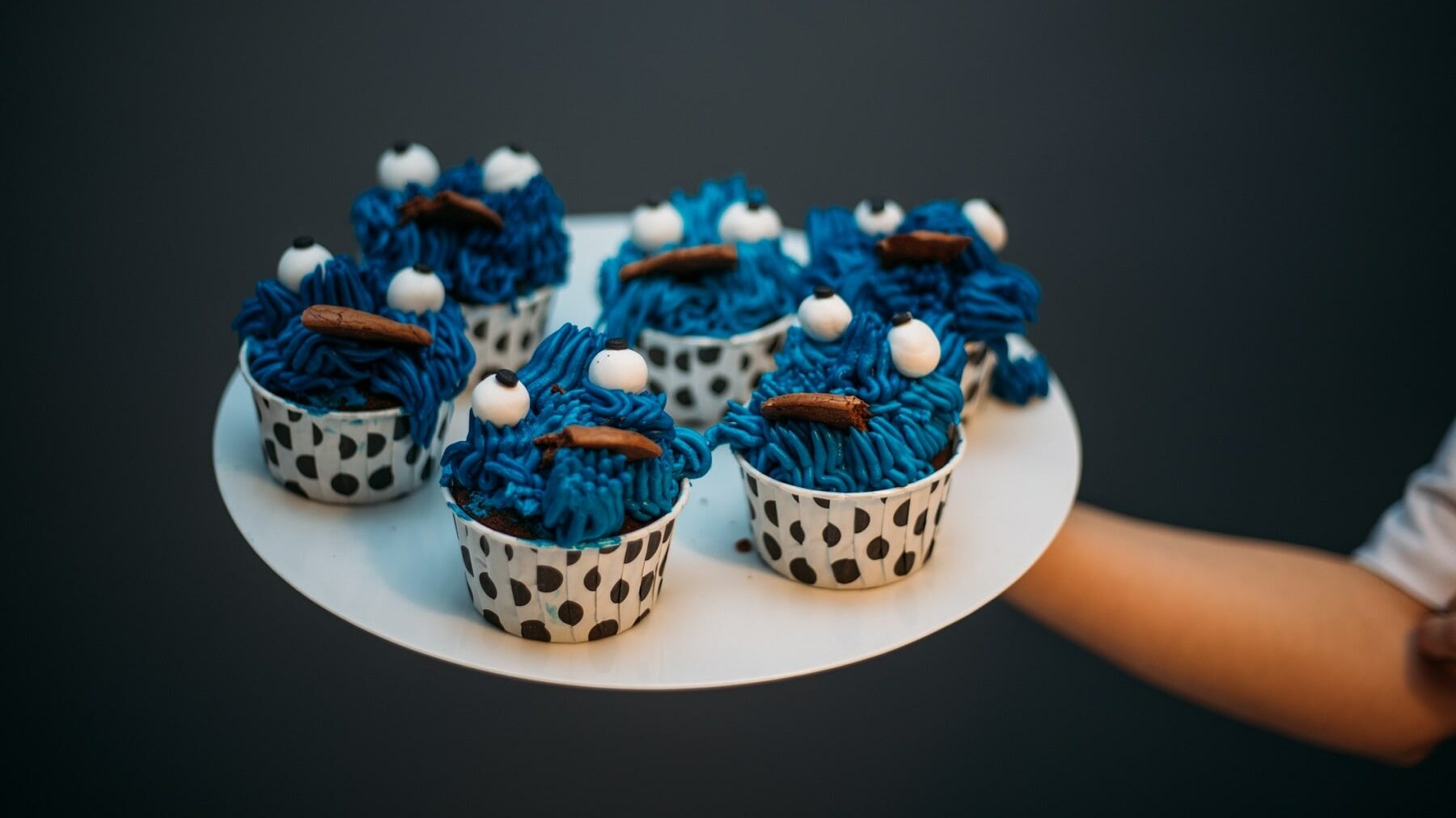 Fünf Krümelmonster-Cupcakes stehen auf einem weißen Teller und werden vor einen schwarzen Hintergrund gehalten. Die Cupcakes stehen in gepunkteten Muffinformen und in jedem Krümelmonster-Cupcake steckt ein Keks.