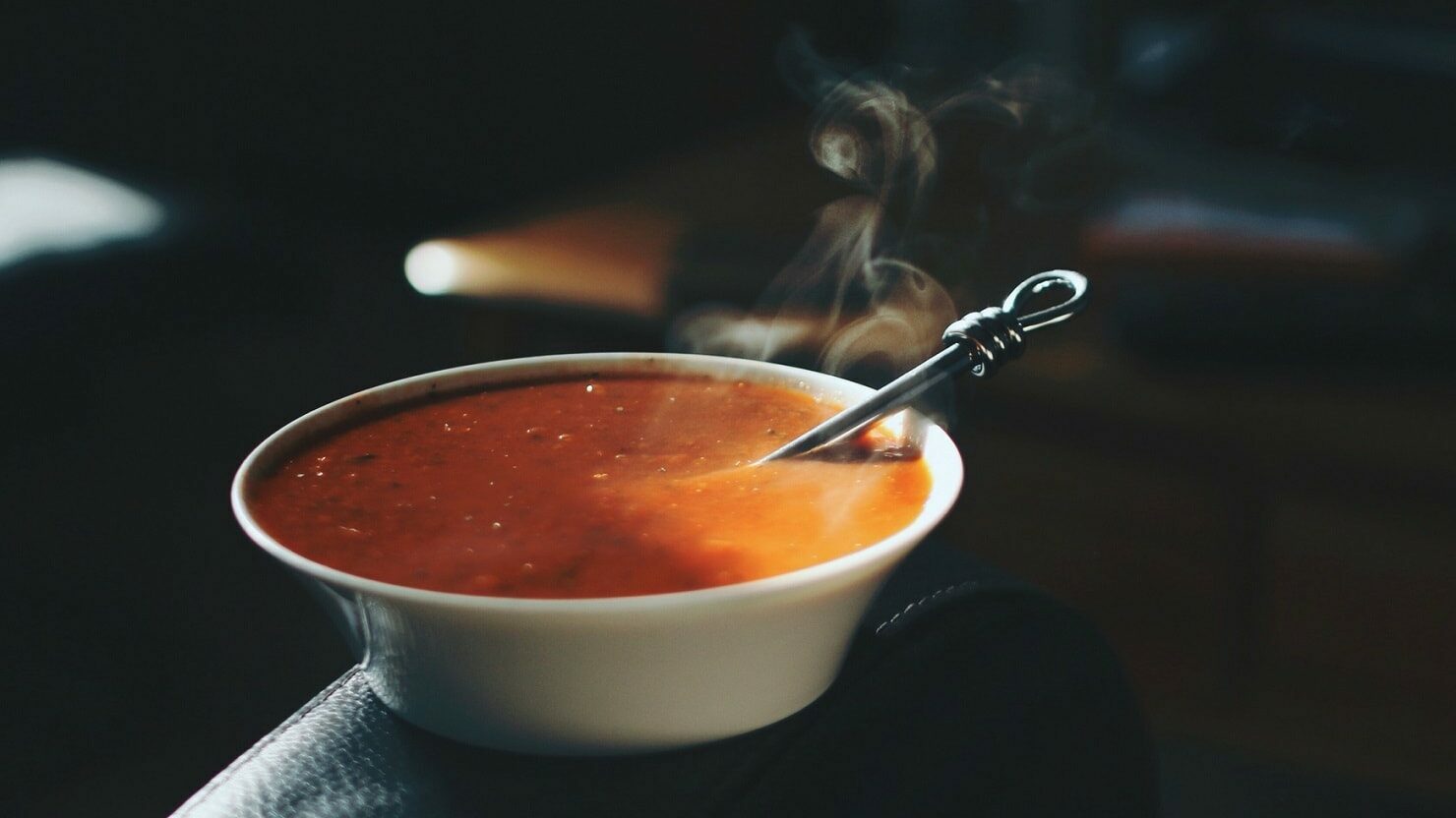Ein Teller mit dampfender, pinkanter Tomatensuppe steht auf der Lehne eines Stuhls in einem dunklen Büro. In der Suppe liegt ein Löffel.