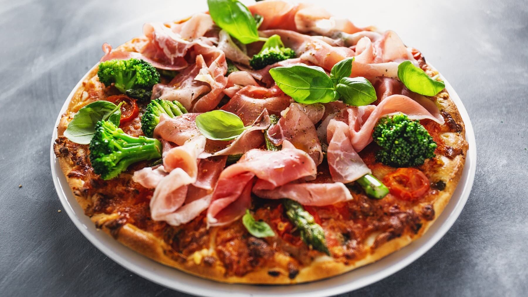 Eine Pizza mit Schinken und Brokkoli steht auf einem weißen Teller auf einem grauen Tisch. Die Pizza ist angerichtet mit frischen Basilikumblättern.