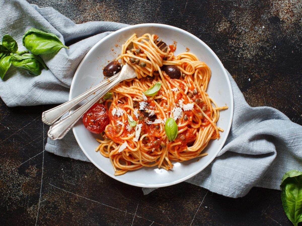 Spaghetti alla puttanesca auf einem Teller mit Besteck. Daneben Basilikum und Parmesan