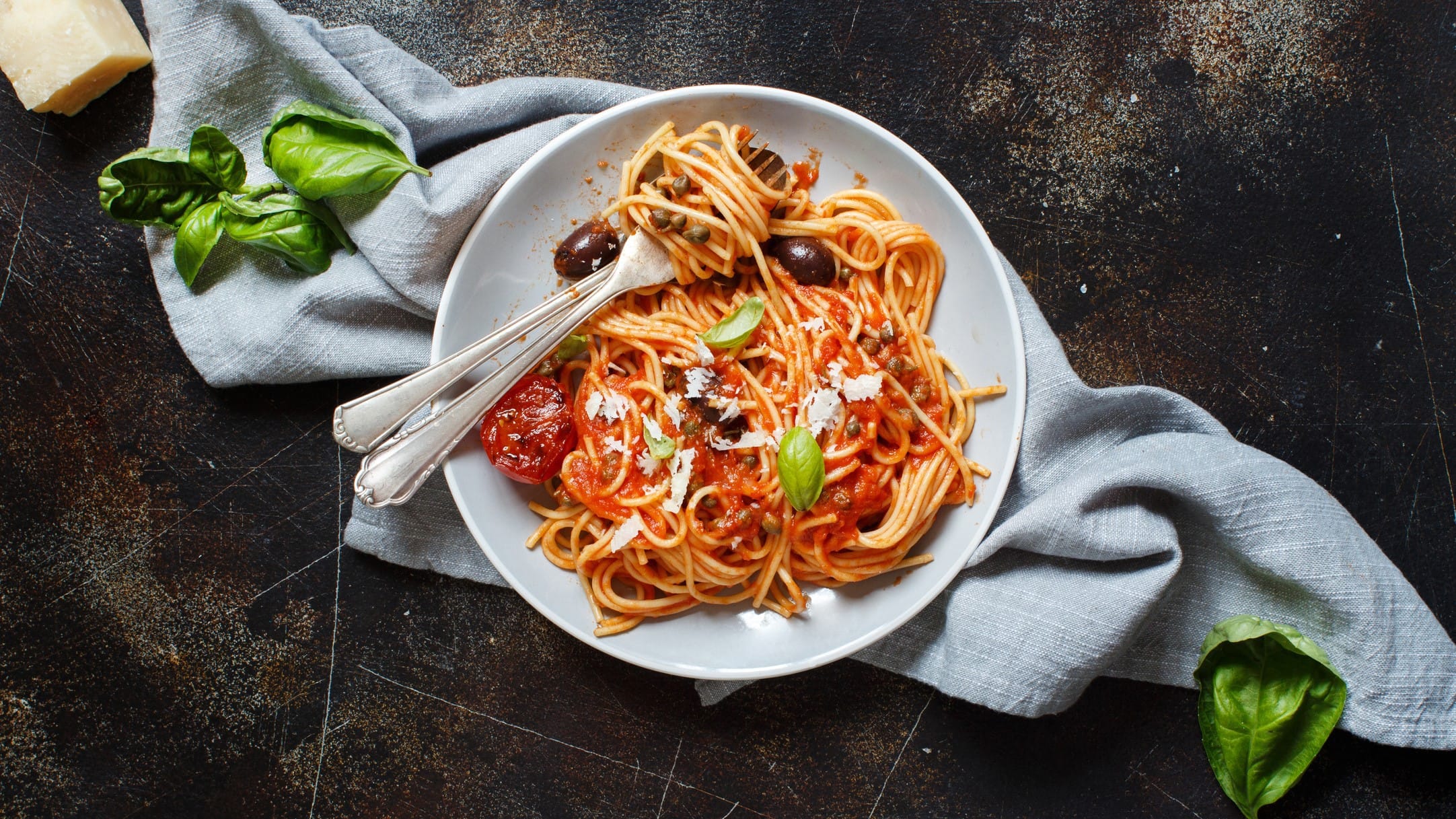 Spaghetti alla puttanesca auf einem Teller mit Besteck. Daneben Basilikum und Parmesan