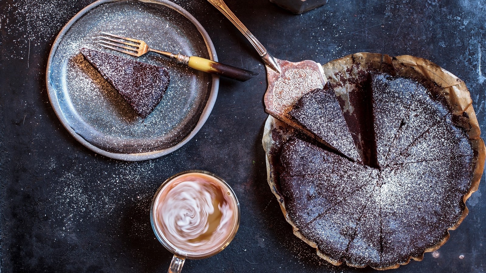 Eine ganze Torta Barozzi mit Puerzucker bestreut und in Stücke geschnitten neben einem Teller mit einem Stück des Kuchens und einer Tasse mit Kaffee, von oben vor dunklem Hintergrund.