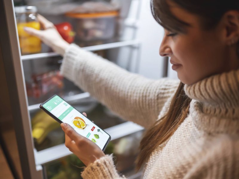 Eine dunkelhaarige Frau steht am geöffneten Kühlschrank und hält ein Smartphone in der Hand, auf dem eine Shopping-App geöffnet ist. NoWaste