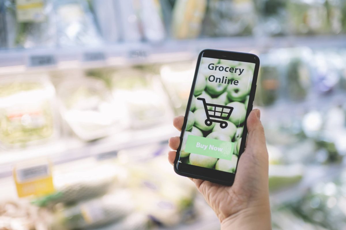 Knuspr: Eine Hand hält ein Handy, auf dem eine App für einen Online-Supermarkt geöffnet ist. Im Hintergrund ist unscharf ein Supermarktregal zu erkennen.